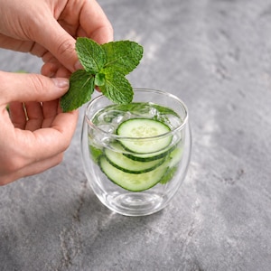 May include: A clear double-walled glass tumbler filled with ice, water, and several thin, round slices of green cucumber. A fresh sprig of bright green mint leaves is positioned above the glass, adding a vibrant herbal element to the drink.
