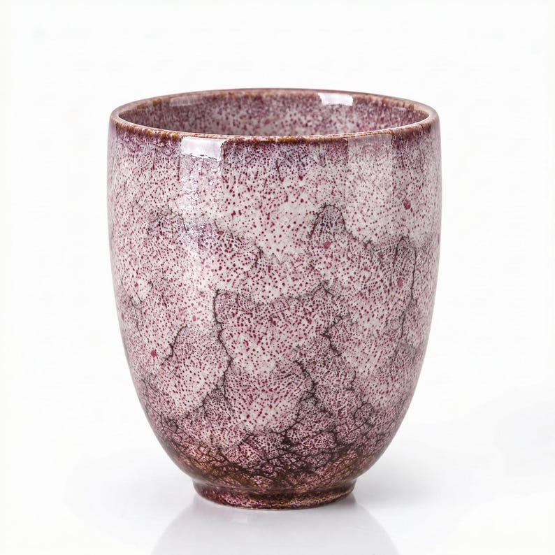 Pode incluir: Uma ch&aacute;vena de cer&acirc;mica com um esmalte salpicado em tons de magenta profundo e branco. A superf&iacute;cie apresenta um padr&atilde;o de fissuras distinto em toda a pe&ccedil;a, com tons castanho-p&uacute;rpura mais escuros concentrados na base e um rebordo arredondado.