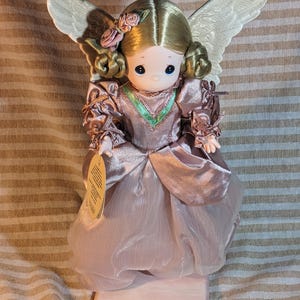 Puede incluir: Una muñeca ángel Precious Moments con cabello rubio, mejillas rosadas y ojos azules. La muñeca viste un vestido rosa con ribete verde y tiene alas blancas. Una etiqueta cuelga de la mano de la muñeca. La muñeca está sobre un soporte rosa.