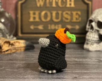 May include: A black crocheted crow wearing an orange and green crocheted pumpkin hat. The crow is sitting on a wooden surface in front of a sign that reads "Witch House 1662".