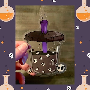 May include: A clear plastic cup with a brown lid and a purple straw. The cup is filled with a dark liquid and has white skull-shaped boba pearls.