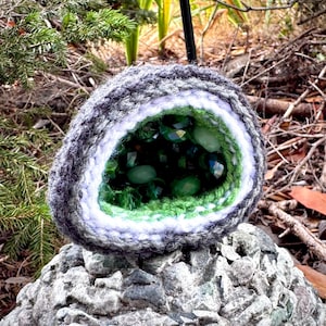 May include: A crocheted geode-shaped object featuring a dark gray outer layer, a white middle ring, and a bright green interior filled with dark green faceted beads. The structure mimics a natural rock formation with a hollow, crystal-like center.