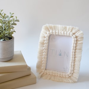 May include: Cream-colored macrame picture frame with a rectangular opening. The frame has a textured, fringed border. The frame is next to a small potted plant and a stack of books. The text "TIAN Design & Macrame" is visible inside the frame.