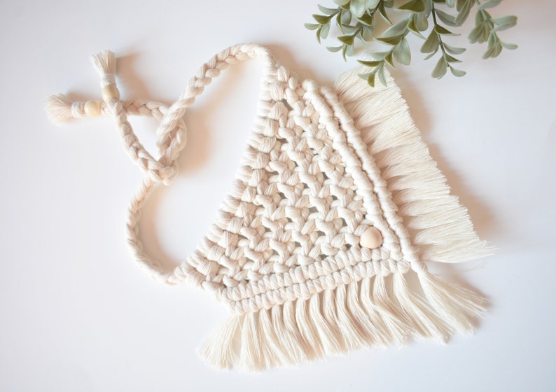 May include: A white macrame wall hanging with a triangular shape and fringe. The hanging has wooden beads woven into the design.