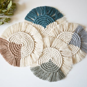 May include: Set of four round macrame coasters with fringe. The coasters are made with natural cotton cord and feature a variety of colors, including beige, gray, teal, and green.