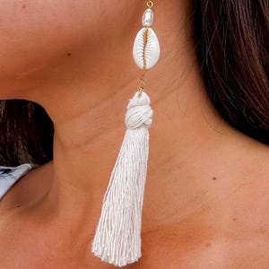 May include: A single gold earring with a white seashell, a pearl, and a long white tassel. The earring is hanging from a gold hook. The tassel is made of white thread and is knotted at the top.