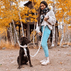 May include: A woman in a white sweater and a black and white scarf walks a brown and white dog on a white rope leash in a forest setting. The trees are bare and the leaves are yellow and orange.