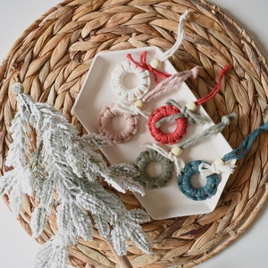 May include: Six handmade macrame ornaments in various colors, including red, white, green, and blue, with wooden beads and tassels. The ornaments are displayed on a white plate on a woven surface.