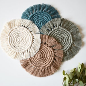 May include: Five handmade macrame coasters in shades of cream, teal, gray, and brown. Each coaster features a circular design with a spiral center and fringed edges. A sprig of green leaves is in the bottom right corner.