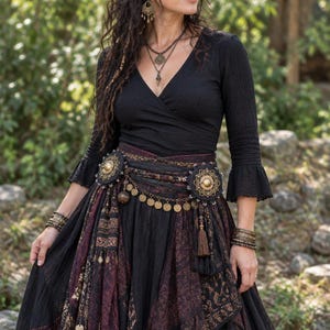 May include: A black long-sleeve wrap top with flared cuffs paired with a layered skirt featuring black and dark maroon patterned fabric panels. The outfit includes a decorative waist belt adorned with large circular gold-toned metal medallions.
