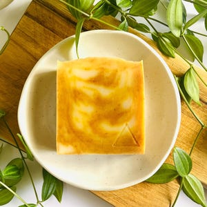 Peut inclure: Un pain de savon carré avec un motif marbré orange et blanc, présentant un petit triangle en relief dans le coin inférieur droit. Le savon repose sur une petite coupelle en céramique blanc cassé, posée sur une planche à découper en bois.