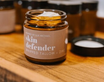 May include: A small, clear glass jar with a brown label that reads "skin defender whipped tallow + zinc." The jar is filled with a white cream and has a black lid. Several similar jars are in the background, all on a wooden surface.
