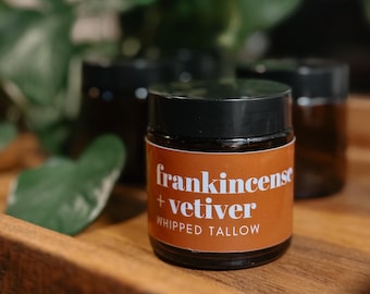 May include: A brown glass jar with a black lid and a white label that reads "frankincense + vetiver WHIPPED TALLOW".