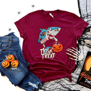May include: A maroon t-shirt with a graphic of a zombie shark holding a pumpkin bucket with the text "Trick or Treat". The shirt is laid out on a white surface with a pair of blue jeans, a skull, and a Halloween party invitation.
