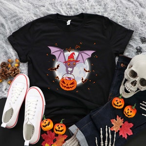 May include: Black t-shirt with a cartoon bat wearing a witch hat and holding a pumpkin. The bat is flying in front of a full moon. There are small bats flying around the moon.