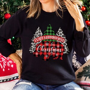 May include: A black sweatshirt with a red and black plaid hippopotamus design. The hippopotamus is surrounded by green and black plaid trees and leopard print trees. The text "I want a hippopotamus for Christmas" is written in white on a red banner.