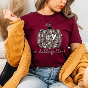 Puede incluir: Una camiseta color burdeos con un dise&ntilde;o de calabaza floral blanco y negro y el texto "hello fall".