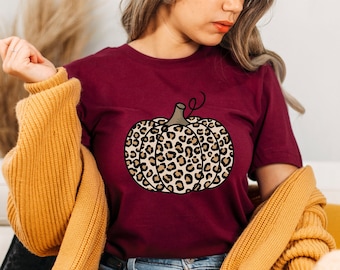 May include: A maroon t-shirt with a leopard print pumpkin graphic. The shirt is being worn by a person with a yellow cardigan.
