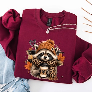 May include: A maroon long-sleeve sweatshirt featuring a graphic of a raccoon wearing an orange knit beanie and a leopard print scarf. The raccoon holds a coffee cup with a heart icon. The design includes orange autumn leaves and a black heart.