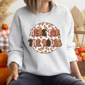 May include: A gray sweatshirt with a white circle design featuring the text "Here for the rolls" in a retro font. The circle is filled with a pattern of brown and pink crescent rolls.