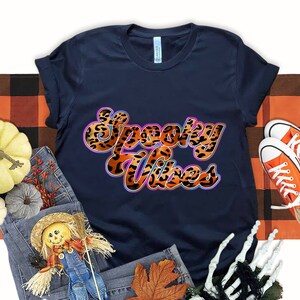May include: A navy blue t-shirt with the text "Spooky Vibes" in orange and black leopard print letters with a purple outline. The shirt is surrounded by fall-themed props, including pumpkins, a scarecrow, and a skeleton hand.