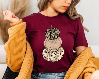 May include: A maroon t-shirt with a stack of three pumpkins. The pumpkins are decorated with a leopard print pattern. The top pumpkin is brown, the middle pumpkin is beige, and the bottom pumpkin is brown.