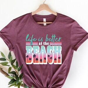 May include: A maroon t-shirt with the text "life is better at the beach" in a retro font with palm trees and a sunset gradient.