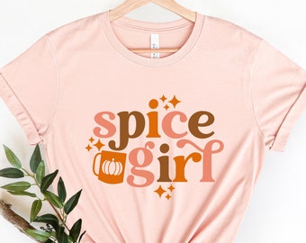 May include: A pink t-shirt with the text "spice girl" in brown and orange letters. A stylized mug with a pumpkin inside is to the left of the text. The mug is orange and brown. There are small white stars around the text.