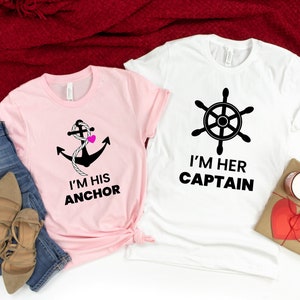Puede incluir: Dos camisetas blancas con texto negro y gráficos. Una camiseta dice "Soy su ancla" con un ancla y un corazón rosa. La otra camiseta dice "Soy su capitán" con un timón.