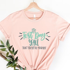 May include: A light pink t-shirt with the text "It's Test Day Y'all Don't Stress! Do Your Best" in green and black lettering.