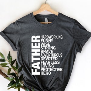 May include: A dark gray t-shirt with the word "FATHER" spelled vertically in white. The word is surrounded by a list of positive adjectives describing a father, including "hardworking", "funny", "wise", "strong", "brave", "adventurous", "devoted", "fearless", "loving", "protective", and "hero".