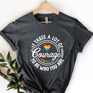 May include: A dark gray t-shirt with a white circle graphic that says "It takes a lot of courage to be who you are". The graphic also includes a rainbow heart.