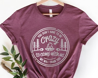 May include: A maroon t-shirt with a silver graphic that says "You don't have to be crazy to camp with us. We will train you." The graphic is a circle with a campfire, trees, and the text inside.