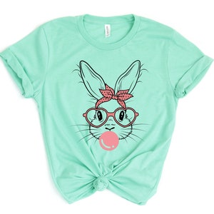 May include: A mint green t-shirt with a graphic of a bunny wearing heart-shaped glasses and a red bandana with white polka dots. The bunny is blowing a pink bubble.