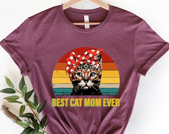 May include: A maroon t-shirt with a retro-style graphic of a cat wearing glasses and a red and white polka dot bandana. The text "BEST CAT MOM EVER" is printed in yellow below the graphic.