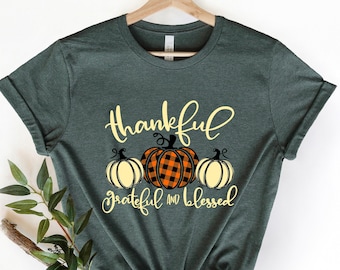 May include: A green t-shirt with the text "thankful grateful and blessed" in yellow lettering. Three pumpkins are centered below the text, one is orange and black plaid, and the other two are white with orange stems.