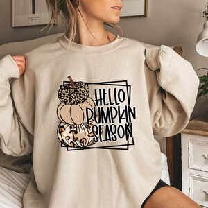 May include: A beige sweatshirt with a graphic design of three pumpkins stacked on top of each other. The pumpkins are decorated with leopard print and the text "HELLO PUMPKIN SEASON" is written in black.