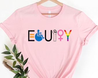 May include: Pink t-shirt with the word "EQUITY" spelled out in black letters. Each letter is decorated with a different symbol representing equality: a wheelchair, a rainbow, a fist, and a female symbol.