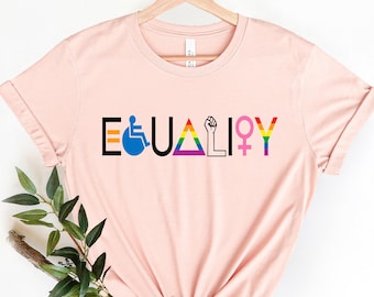 May include: A pink t-shirt with the word "EQUALITY" printed on it in black letters. The letters are replaced with symbols representing different groups, including a wheelchair, a rainbow triangle, a raised fist, and a female symbol.