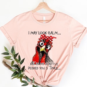 May include: A pink t-shirt with a graphic of a red and black chicken wearing a red bandana with white polka dots and red glasses. The text on the shirt reads "I may look calm... But in my head I've pecked you 3 times..."