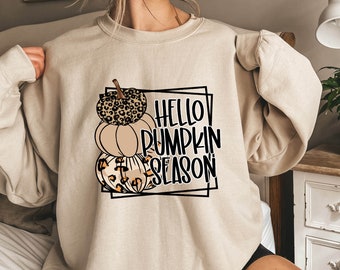 May include: A beige sweatshirt with a graphic design of three pumpkins stacked on top of each other. The pumpkins are decorated with leopard print and the text "HELLO PUMPKIN SEASON" is written in black.