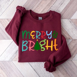 Puede incluir: Una sudadera color burdeos con el texto "Merry & Bright" en letras de colores. Un pequeño árbol de Navidad verde con una estrella amarilla se encuentra entre las palabras.