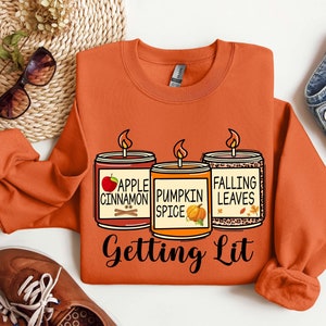 Puede incluir: Sudadera naranja con un gráfico de tres velas con aromas otoñales: manzana canela, especias de calabaza y hojas caídas. El texto "Getting Lit" está debajo de las velas.