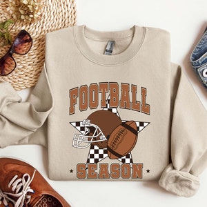 Puede incluir: Una sudadera beige con un diseño gráfico marrón y blanco que presenta un casco de fútbol y el texto "FOOTBALL SEASON".