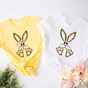 May include: Two t-shirts with a cute cartoon bunny wearing a floral headband and leopard print. The bunny has pink cheeks and black eyes. One shirt is yellow and the other is white.