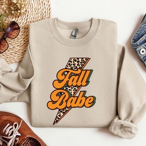 Puede incluir: Una sudadera de color beige con un gráfico de un rayo con un diseño de estampado de leopardo. El texto "Fall Babe" está escrito en letras naranjas y verdes sobre el rayo.