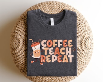 Puede incluir: Una camiseta gris oscura con una taza de café de dibujos animados y el texto "Coffee Teach Repeat" en letras naranja y marrón.
