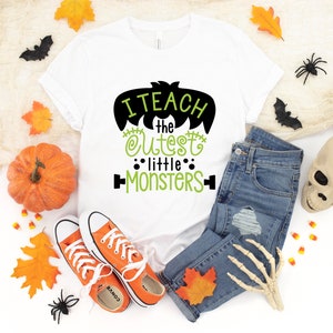 May include: White t-shirt with a black and green graphic that says "I teach the cutest little monsters". The shirt is surrounded by Halloween decorations, including pumpkins, leaves, spiders, and a skull.