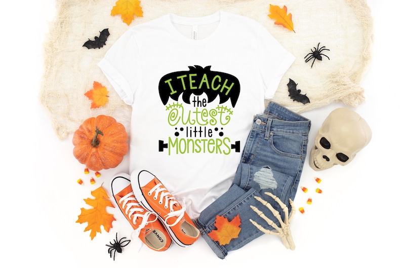 May include: White t-shirt with a black and green graphic that says "I teach the cutest little monsters". The shirt is surrounded by Halloween decorations, including pumpkins, leaves, spiders, and a skull.