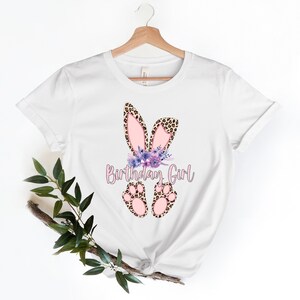 K&ouml;nnte beinhalten: Wei&szlig;es T-Shirt mit einem pinkfarbenen und braunen Hasenohren-Design und dem Text "Birthday Girl".